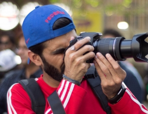 man in red and white track jacket wearing blue baseball cap and black backpack taking photo using bridge camera near people during daytime thumbnail